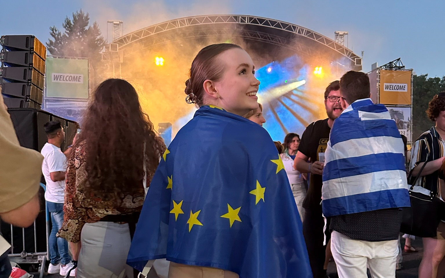 Etualalla nuori nainen EU-lippu harteilla hymyillen, katsoo oikealle, takana seisovia ihmisiä ja suuri esityslava, jolta loistaa keltaisia ja sinisiä värivaloja  savukoneen sumun läpi.
